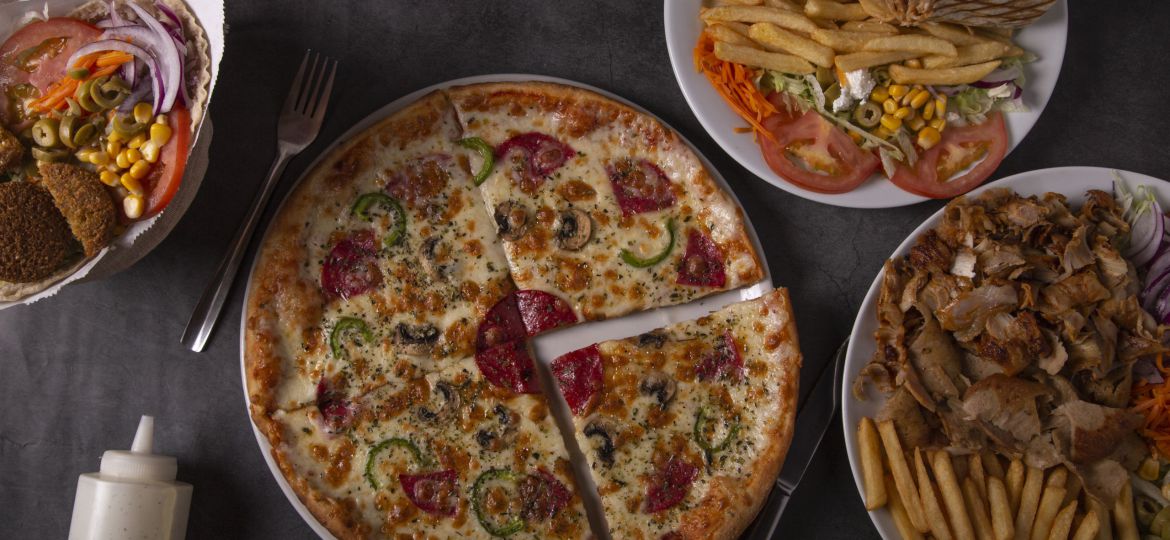 Top view of table scene with a variety of foods for take out or delivery. Beef and chicken kebab, pepperoni pizza and sauces, including fries.